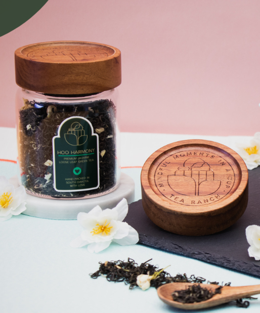 Tea Ranch Hoo Harmony premium loose-leaf jasmine green tea in a reusable glass jar with an acacia wood lid, displayed with loose tea leaves, jasmine blossoms, and the engraved Tea Ranch wooden lid.