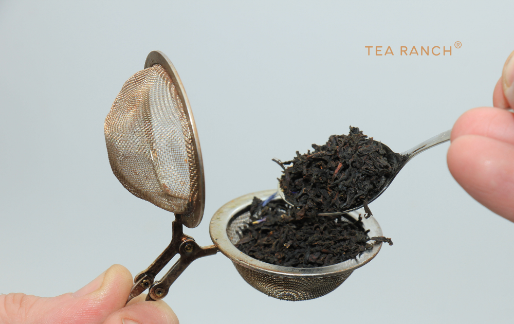 Spoon adding loose-leaf black tea into a reusable stainless steel mesh tea infuser before brewing.
