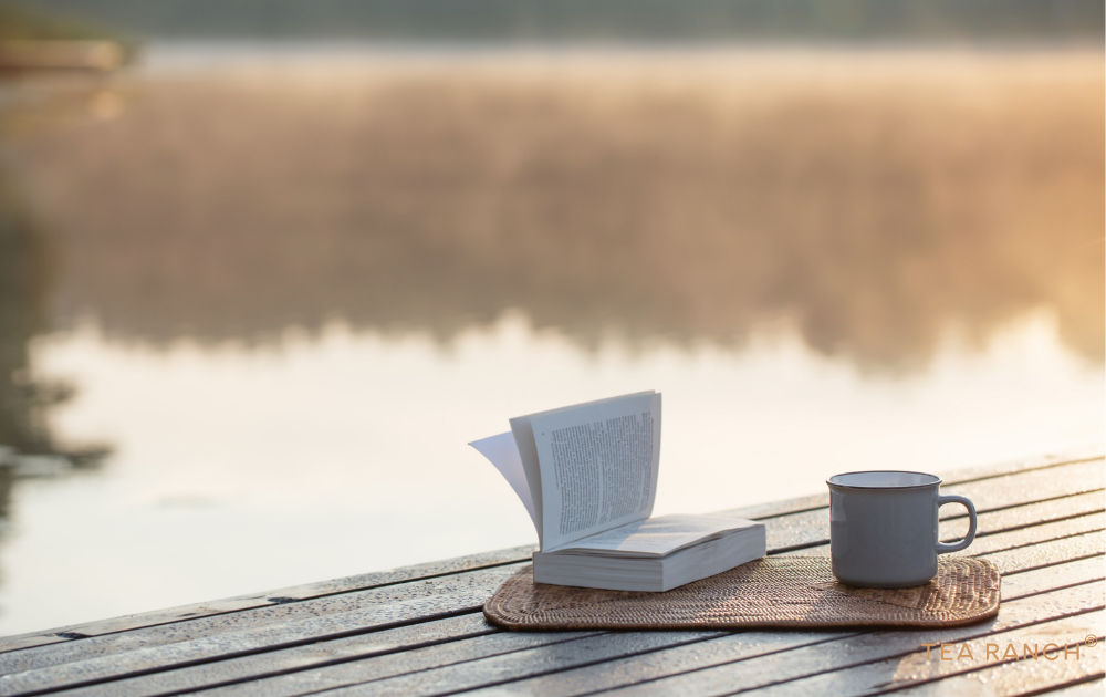 Steaming cup of tea on a wooden dock overlooking a peaceful lake at sunrise. Cup of loose-leaf tea and an open book on a dock overlooking a peaceful lake at sunrise, illustrating the calming effects of spending time near water.
