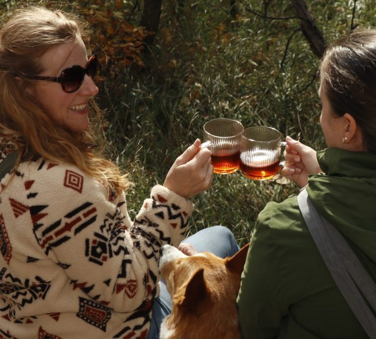 A warm lifestyle photograph of two friends enjoying freshly brewed loose-leaf tea while relaxing together in a woodland setting with their dog. Surrounded by trees and natural grasses, the image captures Tea Ranch's belief that meaningful connections are often found through shared moments in nature. Two women cheerfully clinking their glasses of tea in a forested area, with a dog nearby.